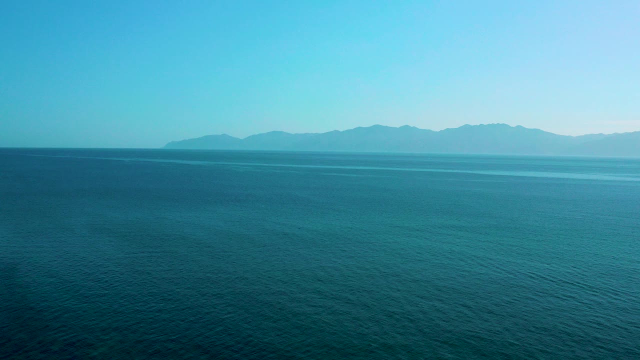 Aerial view over blue ocean toward a hazy mountain range in El Sargento La Ventana, Mexico, with no visible people.