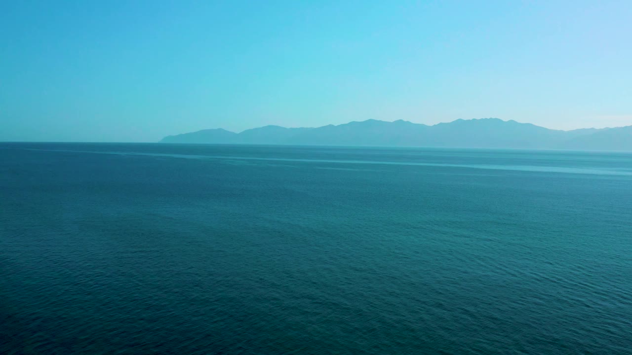 Aerial frame of textured blue sea in El Sargento La Ventana, Mexico, with a long distant mountain range and clear daylight sky, no visible people.