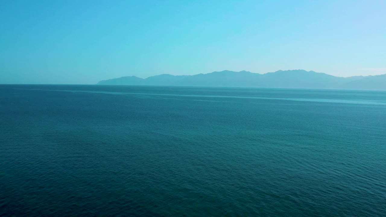 Wide aerial view over deep blue water in El Sargento La Ventana, Mexico, with a narrow lighter band of sea near the mountain horizon and no visible people.
