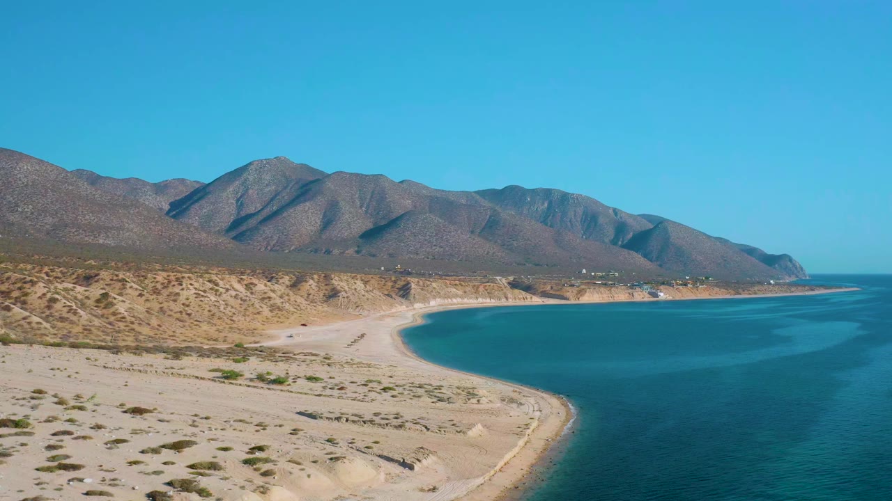Aerial view over El Sargento La Ventana, Mexico, with pale beach in the foreground, blue bay water to the right, brown mountains in the distance, and no visible people.