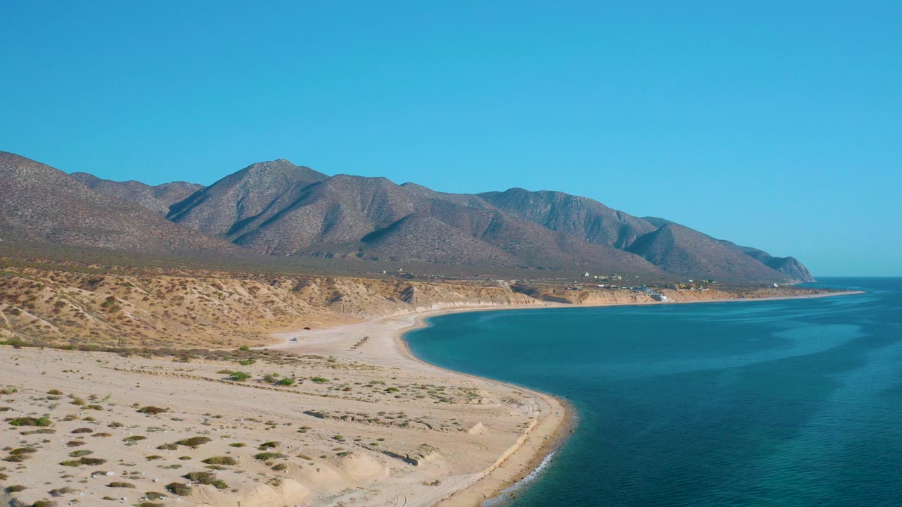 Opening frame of the El Sargento La Ventana coastline with pale beach foreground, turquoise water at right, brown hills beyond, and no visible people.