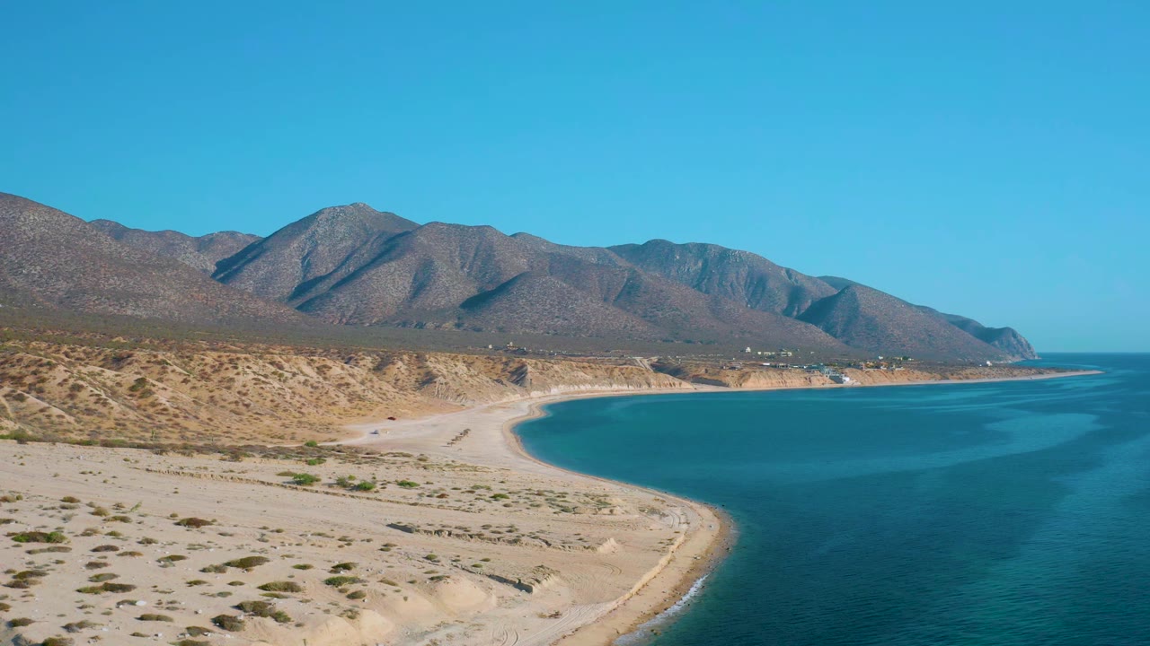 Early frame shows the curved sandy shoreline reaching into the blue bay near El Sargento La Ventana, with arid mountains filling the horizon and no visible people.