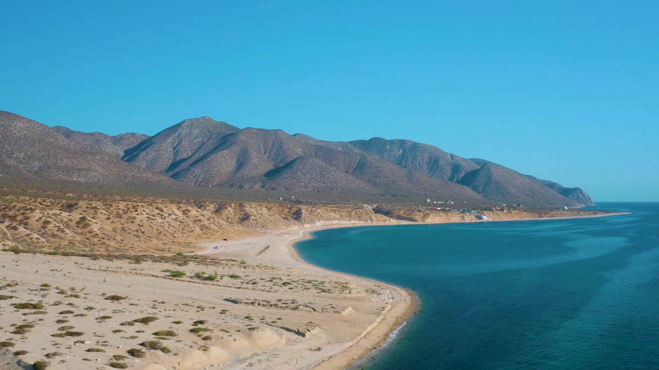 The frame holds a wide view of the Mexico shoreline, with shallow turquoise tones near the beach, deeper water to the right, dry mountain slopes behind, and no visible people.