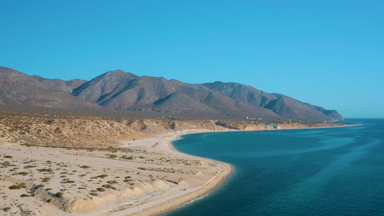 Opening aerial frame over El Sargento La Ventana, Mexico, showing scrubby sand flats, a curved beach, blue water to the right, distant mountains, and no visible people.