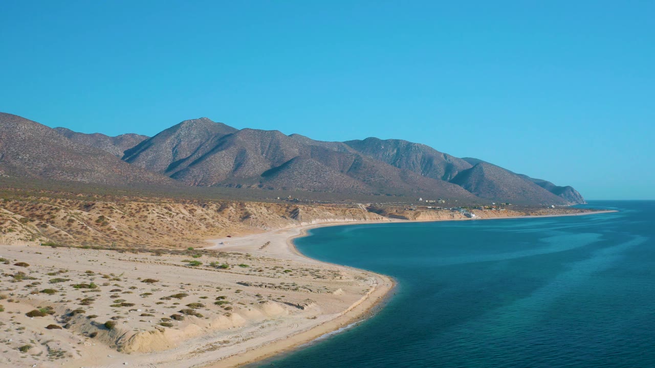 Aerial frame in El Sargento La Ventana, Mexico, showing pale sand, sparse coastal scrub, turquoise water, a low shoreline point, rugged mountains, and no visible people.