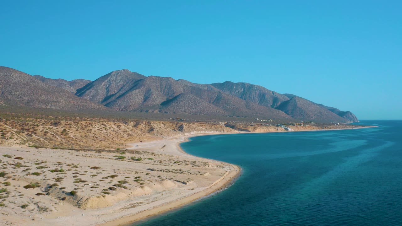 Mid-clip aerial view of El Sargento La Ventana, Mexico, with sandy flats in the foreground, a rounded beach edge, blue sea at right, mountain slopes across the horizon, and no visible people.