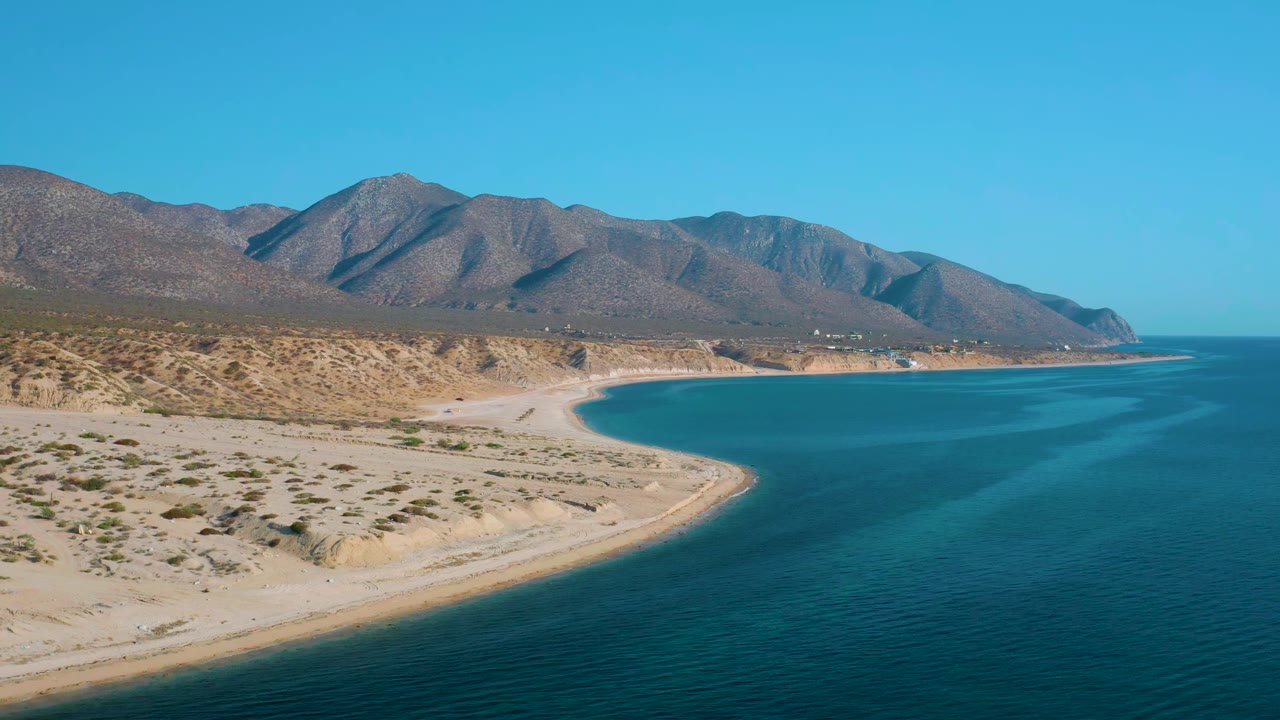 At 0:01, the coastline fills more of the lower frame while the blue bay curves toward distant mountains in El Sargento La Ventana, with no visible people.