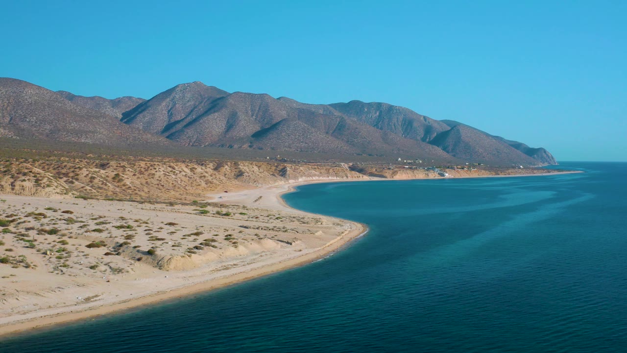 At 0:02, the sandy point and scrubland sit at left while the broad blue water and mountain ridge extend across the frame, with no visible people.