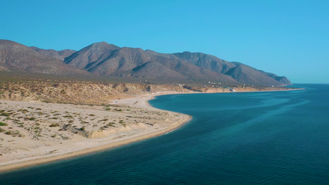 At 0:03, scrub-covered sand occupies the foreground, the shoreline bends through the middle, and calm blue water stretches toward the right, with no visible people.