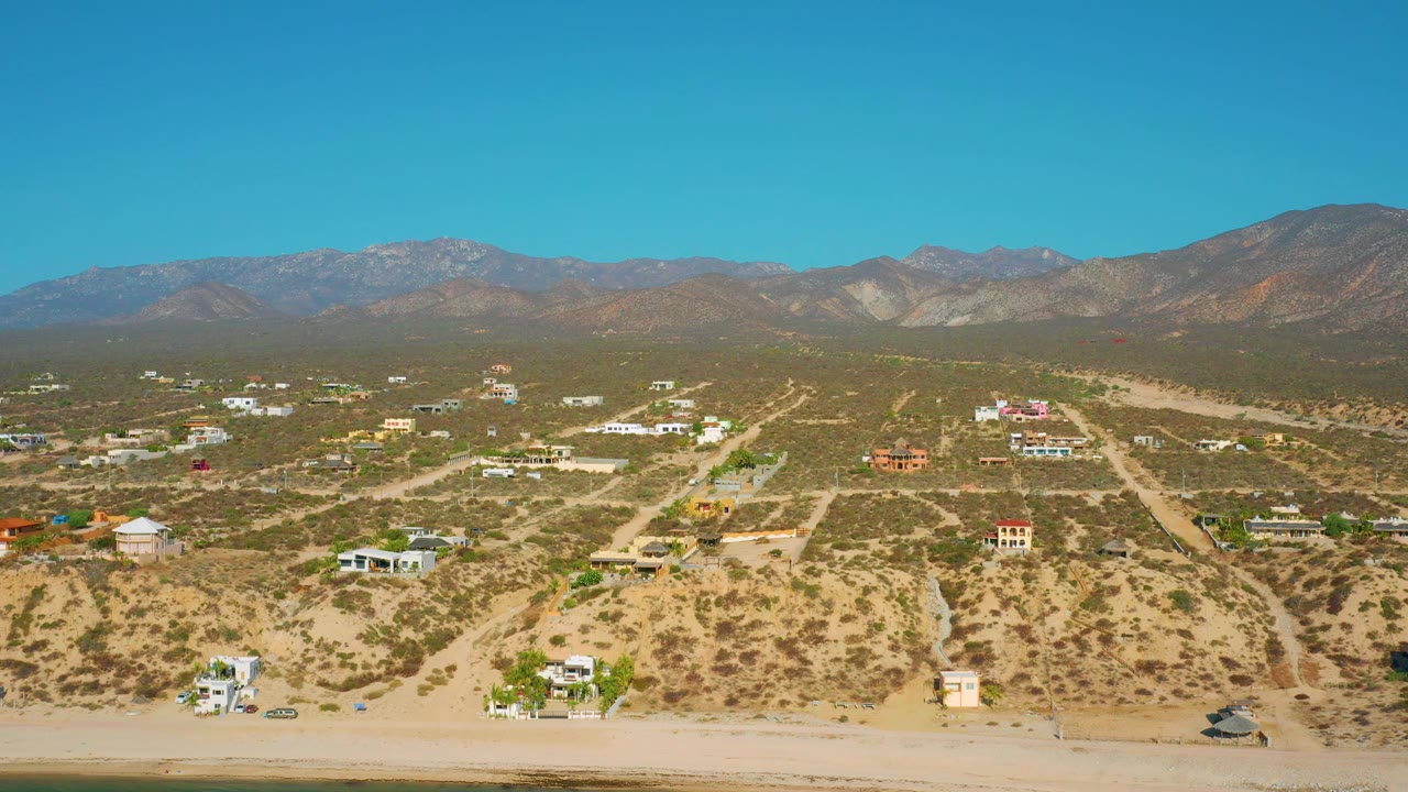 Aerial frame over El Sargento La Ventana showing a clearer cluster of homes in the midground, dusty road lines, dry vegetation, mountains across the horizon, and no visible people.