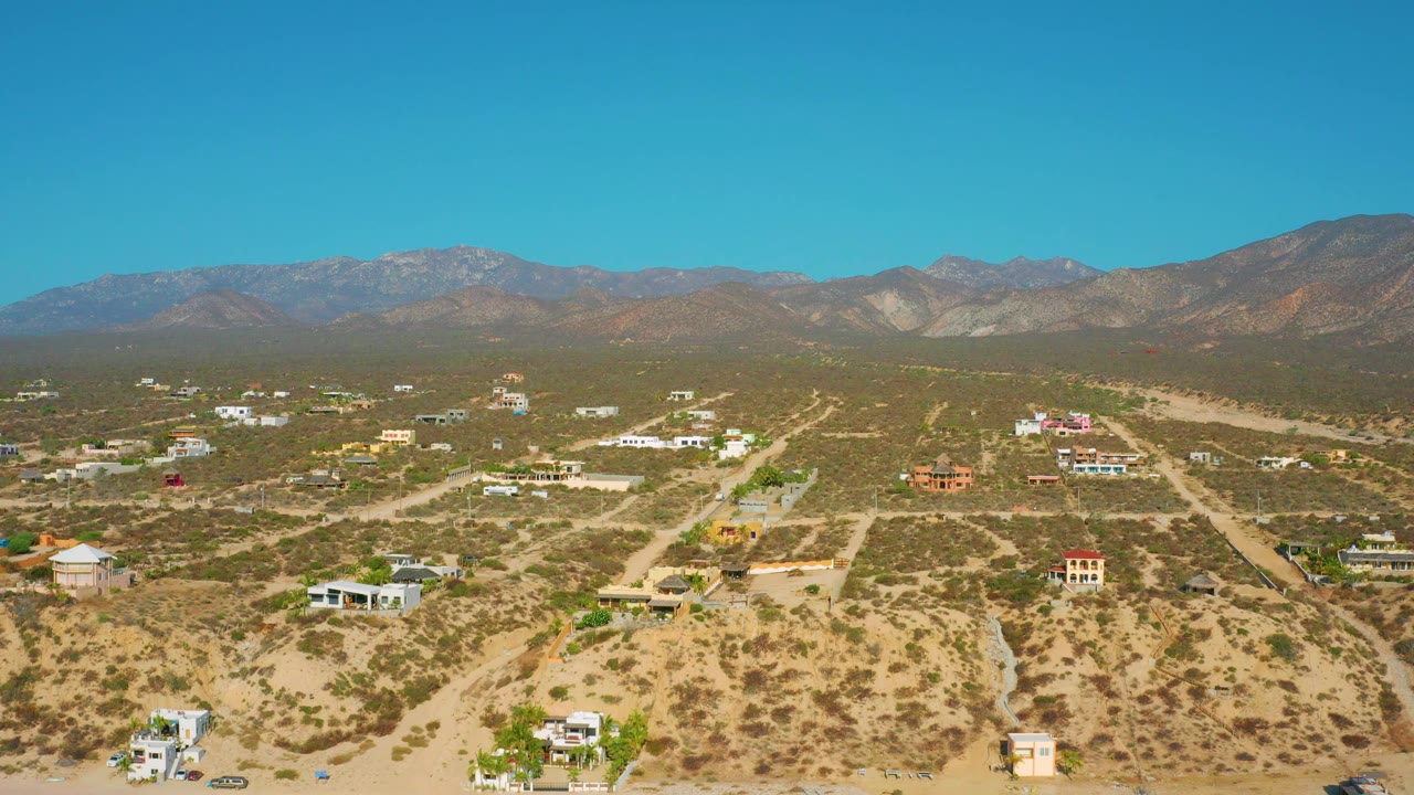 Wide drone-style frame over El Sargento La Ventana, Mexico, with scattered homes across scrubby desert, road cuts through the land, mountains behind, and no visible people.