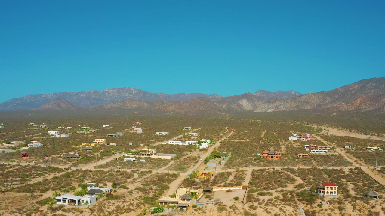 Wide aerial frame of El Sargento La Ventana, Mexico, showing scattered residential buildings across arid lots, a long dirt road at right, hazy mountains, and no visible people.