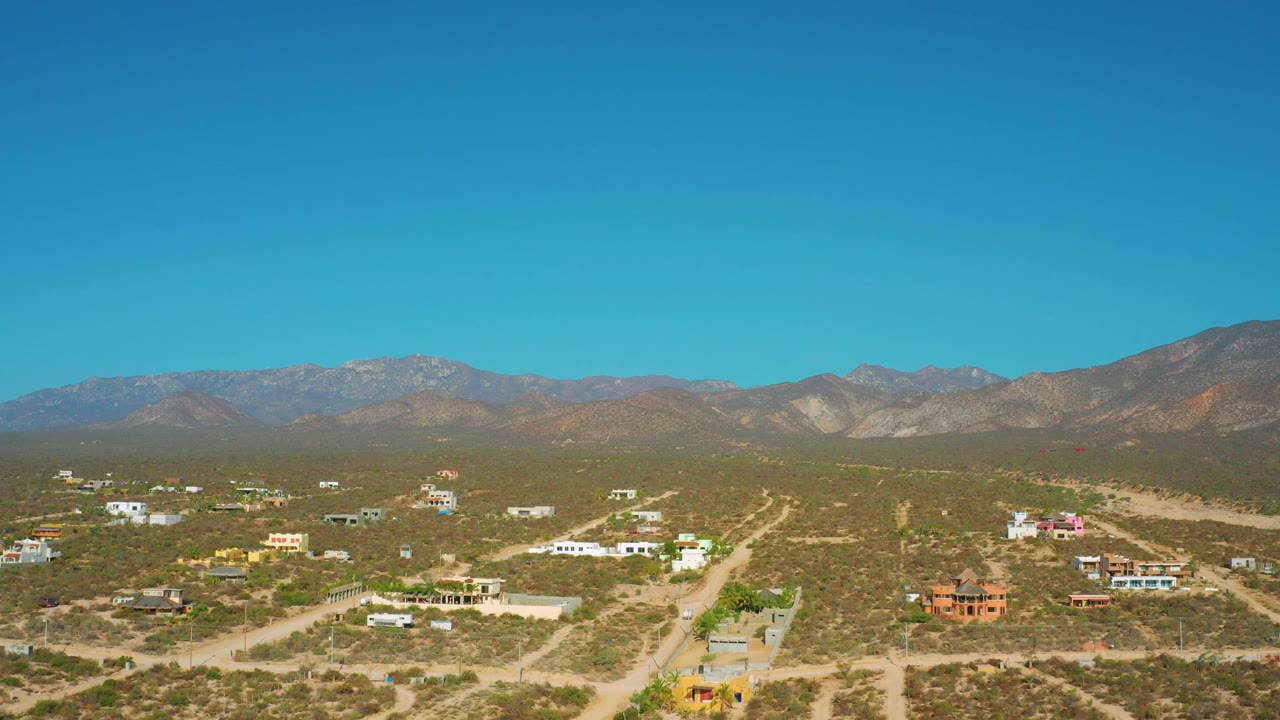 El Sargento La Ventana aerial frame showing scattered white and colored buildings across dry scrubland, a broad mountain horizon, and no visible people.