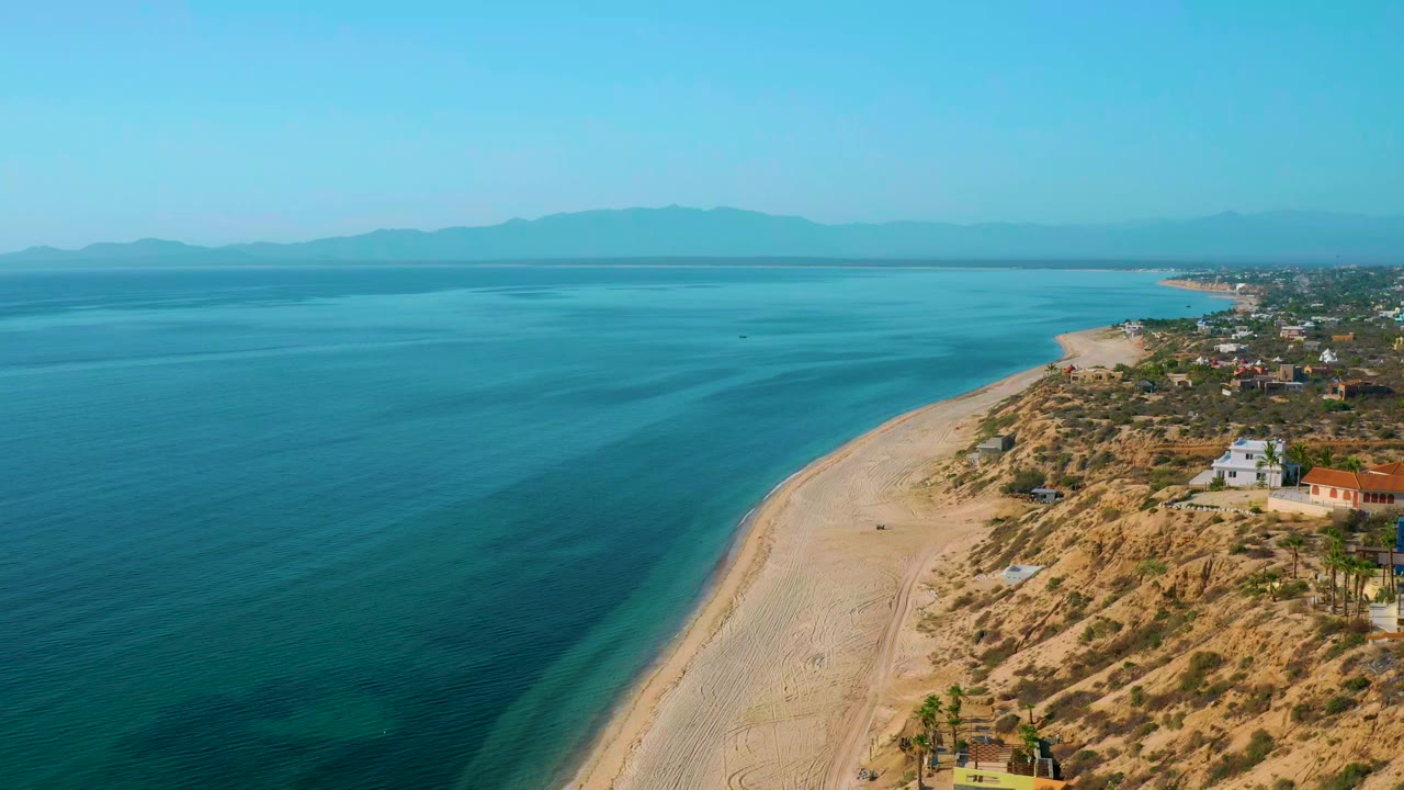 Aerial frame over El Sargento La Ventana, Mexico, showing blue ocean, a pale curving beach, scrub dunes, scattered coastal homes, distant mountains, and no visible people.