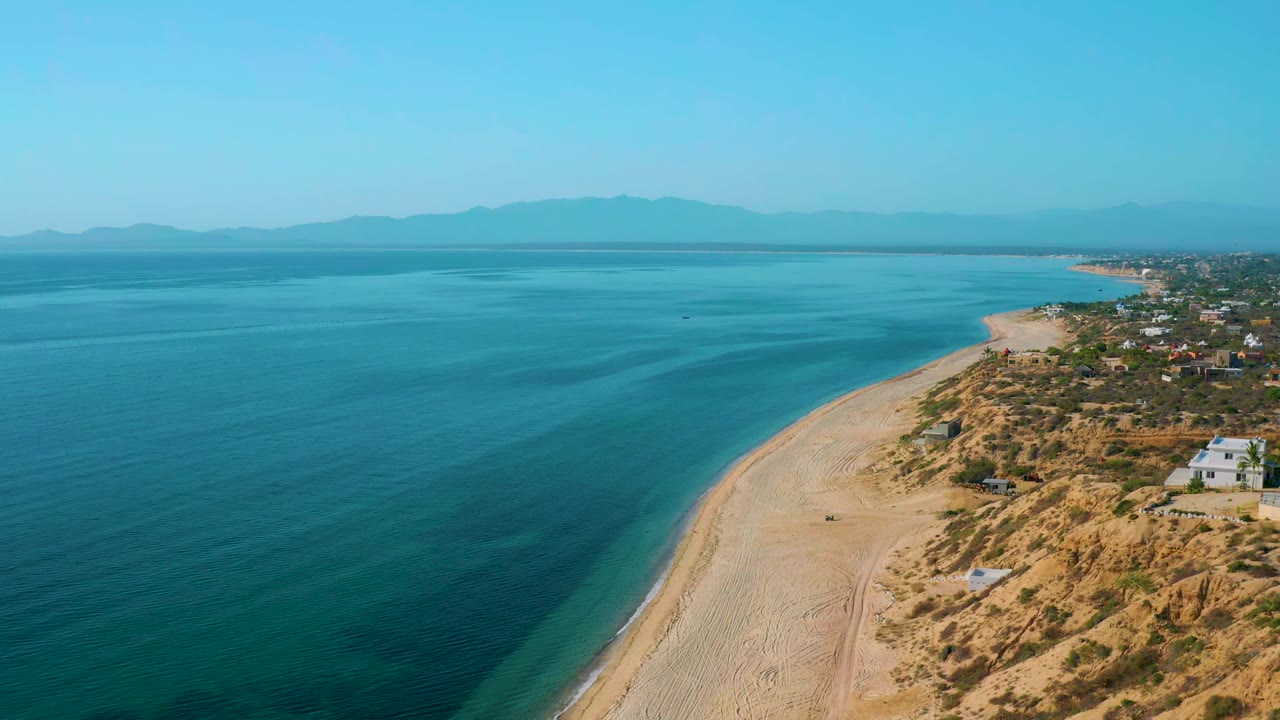 Drone view of a long sandy beach separating blue sea and coastal houses.