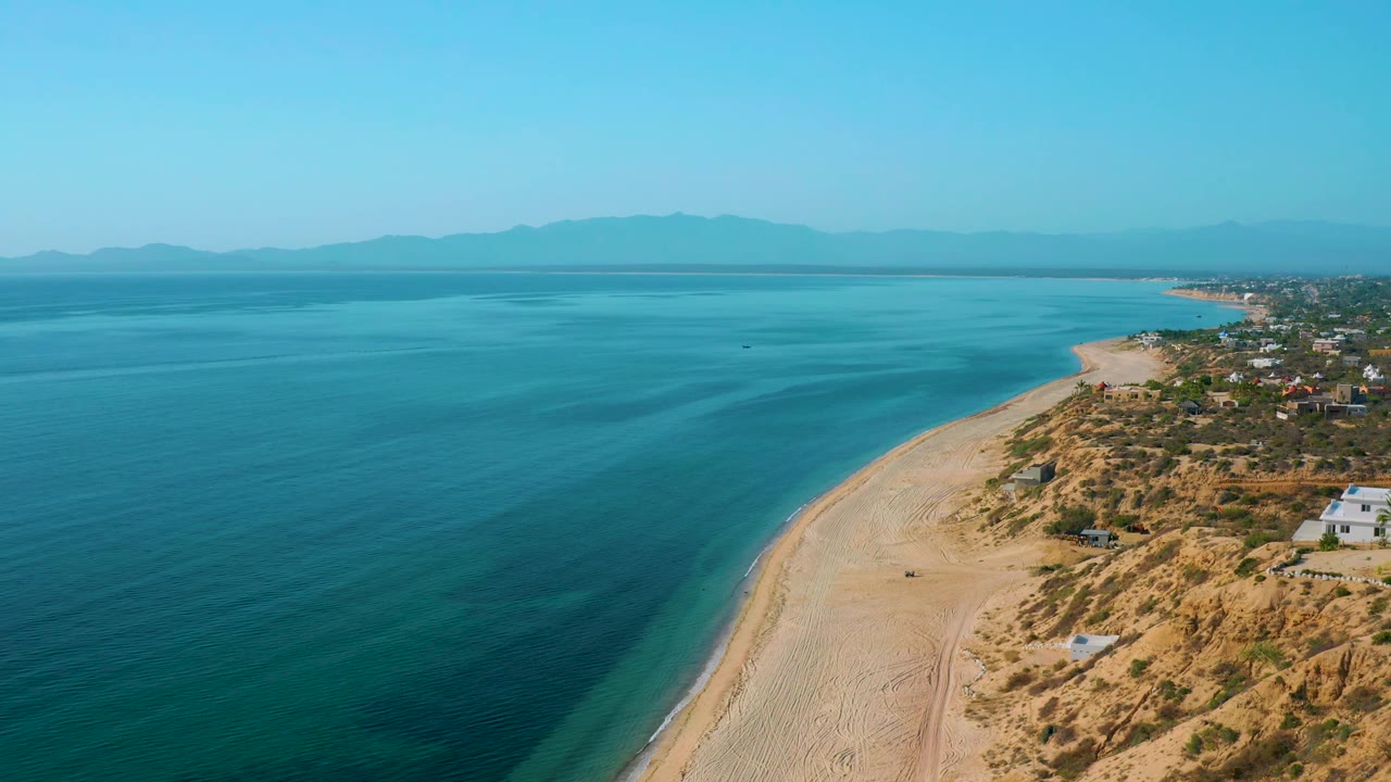 Wide high view over El Sargento La Ventana, Mexico, showing turquoise water across the left and center, a pale beach along the lower right, scrubland and houses near the edge, and no visible people.