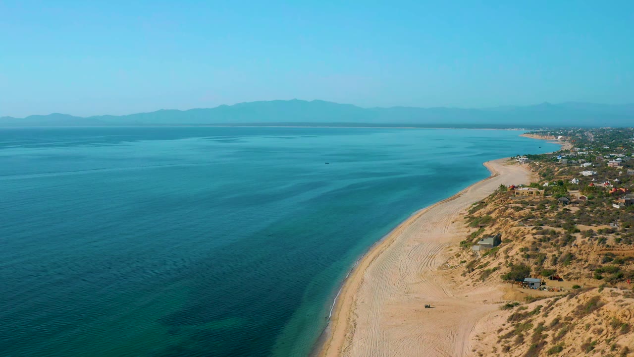 Aerial frame in El Sargento La Ventana, Mexico, looking along the curving beach with turquoise shallows near shore, deeper blue water offshore, hillside homes to the right, and no visible people.