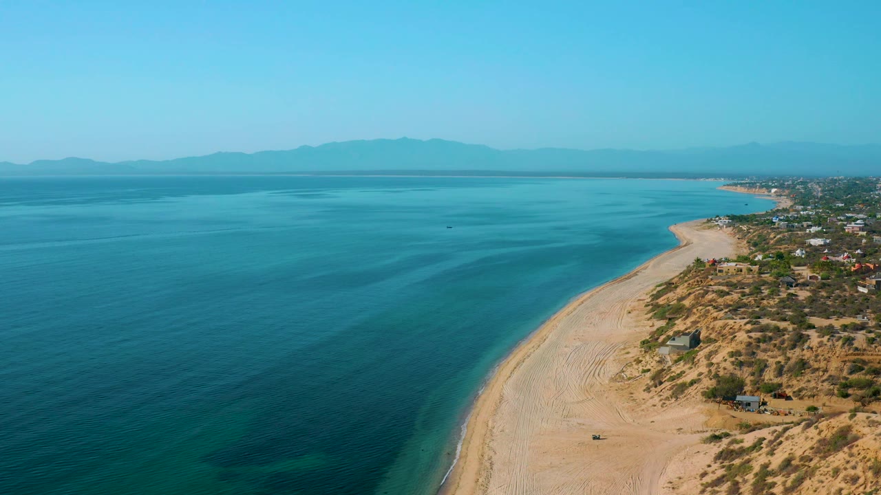 Wide aerial frame over El Sargento La Ventana, Mexico, with a broad expanse of ocean, pale sand running diagonally, clustered coastal buildings at the far right, hazy mountains beyond, and no visible people.