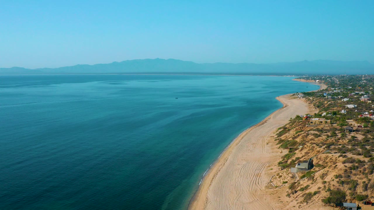 High aerial view over El Sargento La Ventana, Mexico with blue ocean on the left, pale beach curving along the shore, dry scrubland and scattered buildings on the right, and no visible people.