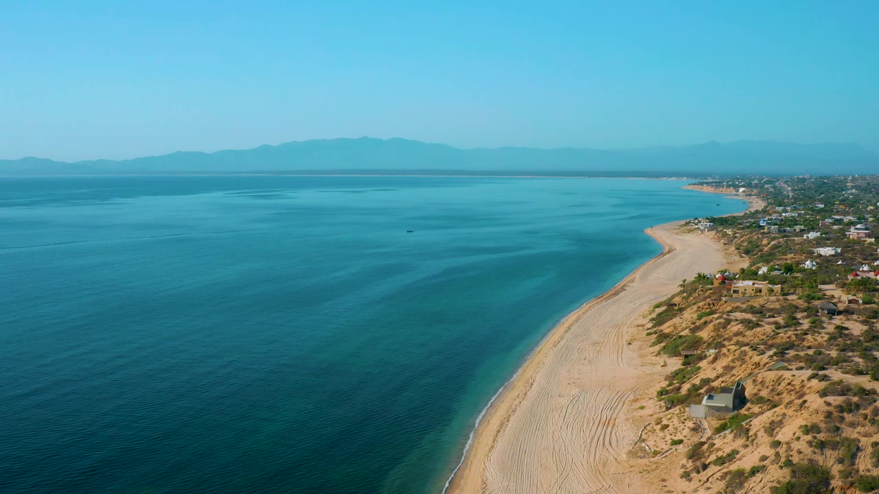 Wide drone view over El Sargento La Ventana, Mexico with open water dominating the left side, a sandy shore running diagonally, coastal homes set back from the beach, and no visible people.