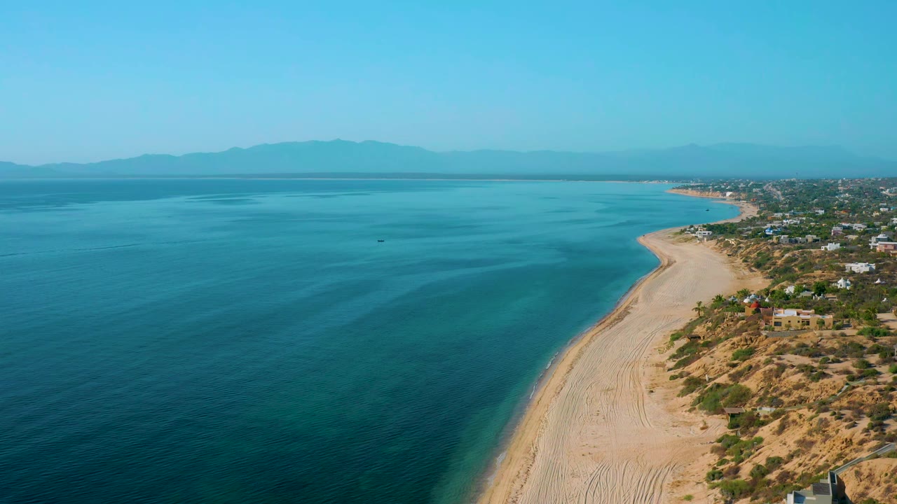 Second aerial frame of El Sargento La Ventana, Mexico, showing turquoise shallows near the beach, deeper blue water offshore, a small dark boat-like speck on the sea, coastal homes on dry terrain, distant mountains, and no visible people.