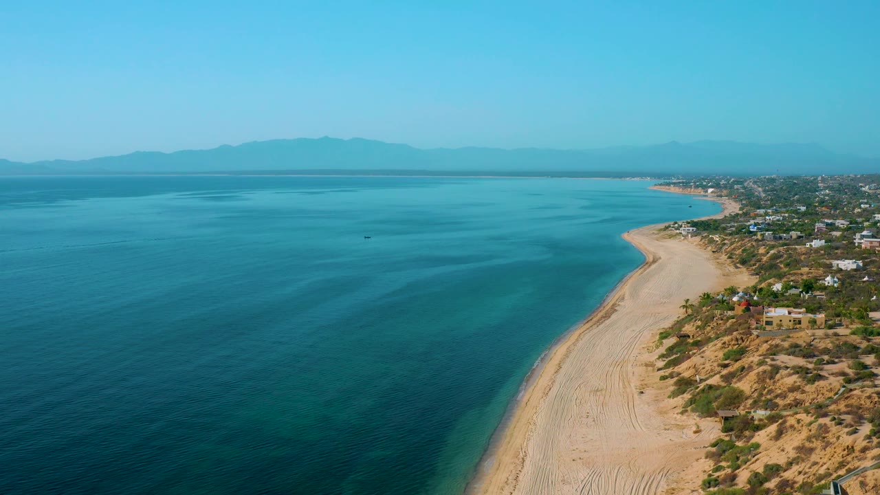 Third aerial frame over El Sargento La Ventana, Mexico, with the beach running from lower right toward the distant coast, blue water spread across the left, small coastal structures on the right, mountain ridges on the horizon, and no visible people.