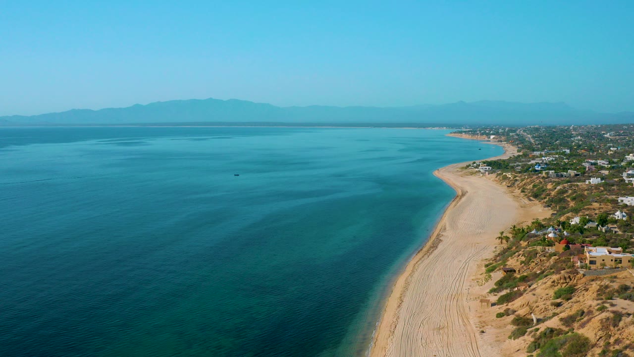 High aerial view of El Sargento La Ventana, Mexico, with deep blue ocean on the left, a pale sand shoreline curving along the right, coastal homes beyond the beach, distant mountains, and no visible people.