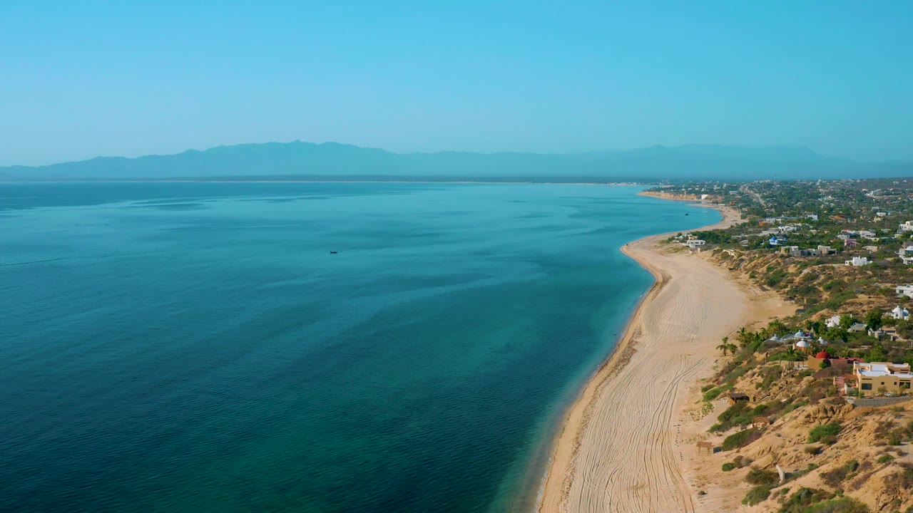 Wide aerial view at El Sargento La Ventana, Mexico, with open blue sea filling most of the frame, a curving sand beach at right, scattered shoreline homes, hazy mountain silhouettes, and no visible people.