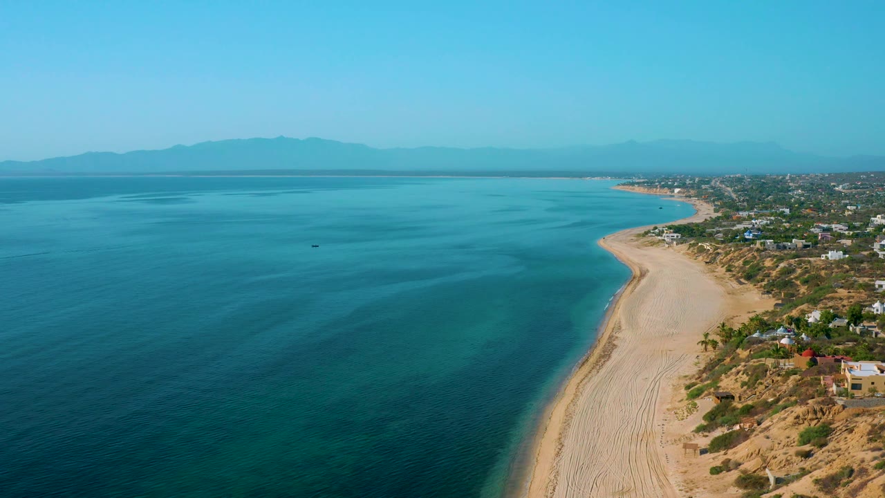 Aerial beach edge with dark blue ocean, pale sand, houses, and mountains.