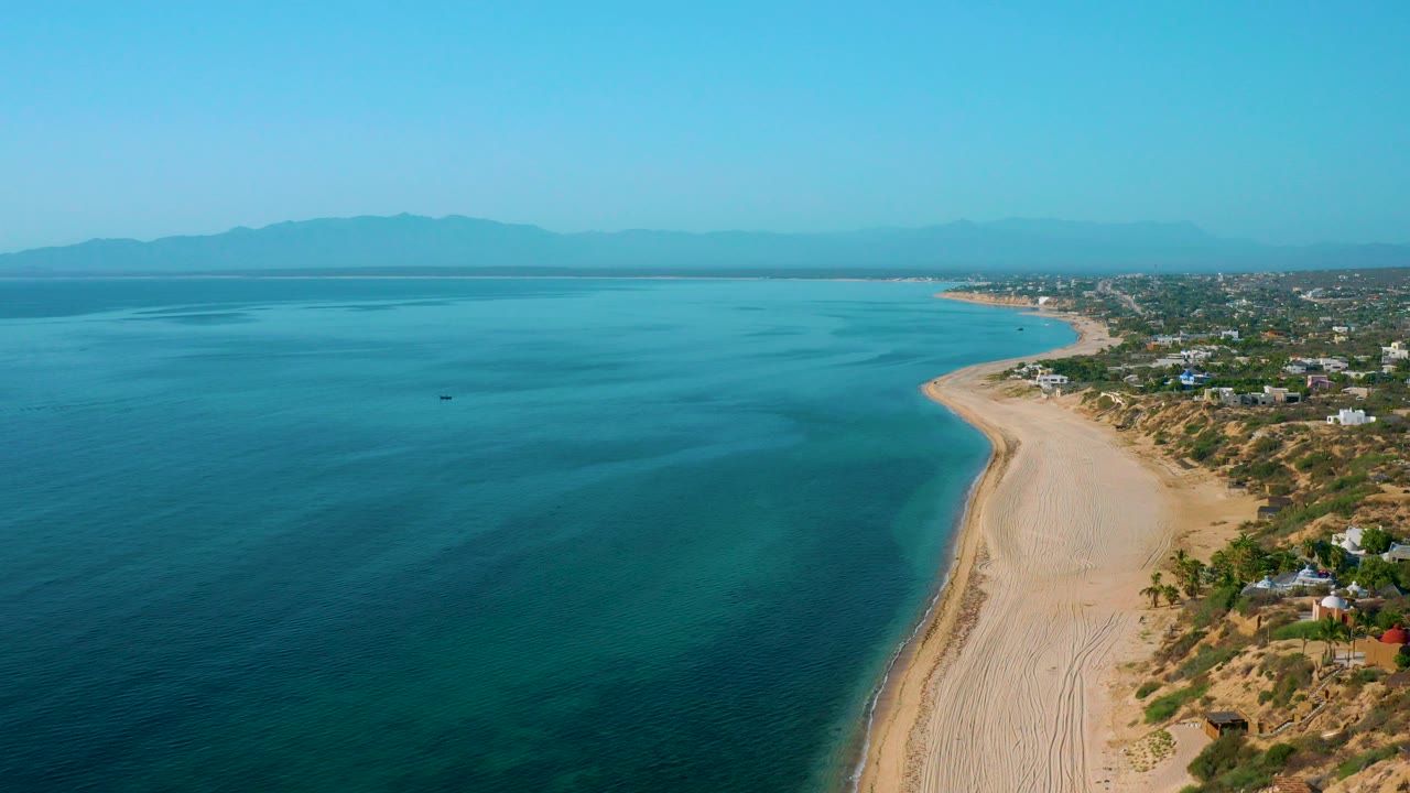 High aerial view over El Sargento La Ventana, Mexico, showing turquoise water on the left, a sandy beach on the right, coastal homes beyond the sand, distant mountains, and no visible people.