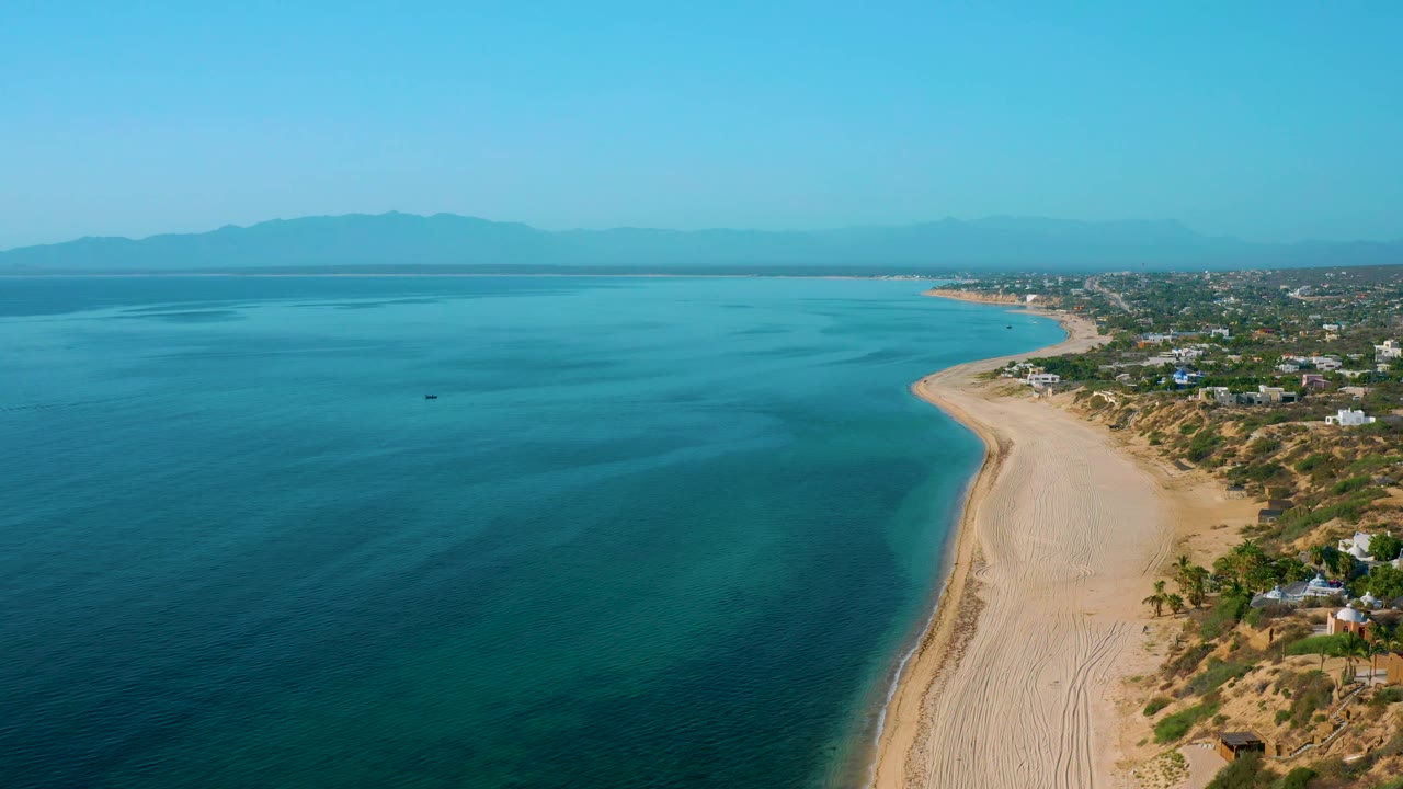 Wide drone view over El Sargento La Ventana, Mexico, with clear shallow water near the beach, deeper blue water offshore, houses lining the coast, distant mountain silhouettes, and no visible people.