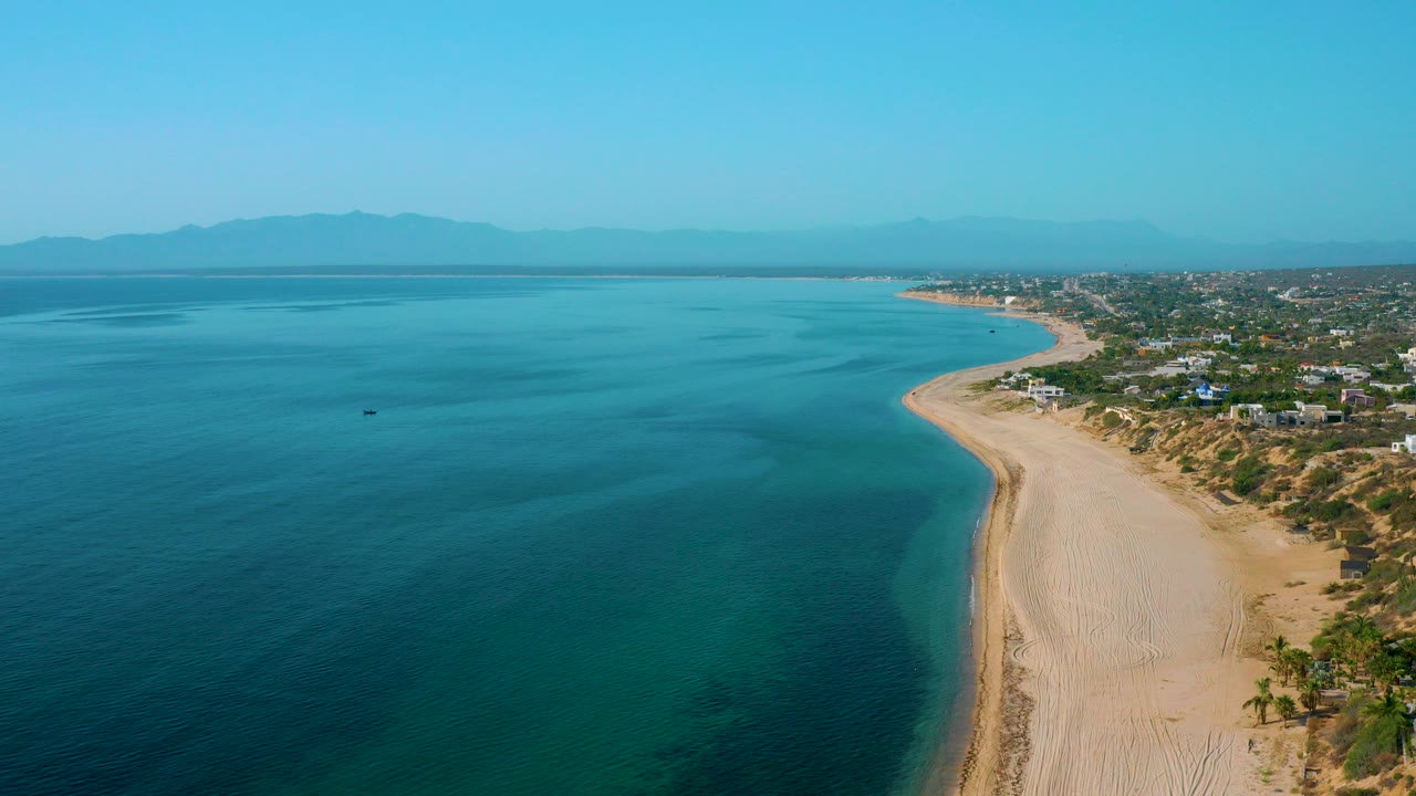 Opening aerial frame over El Sargento La Ventana, Mexico, with deep blue water on the left, a pale sandy beach curving along the right, coastal homes inland, distant mountains, and no visible people.