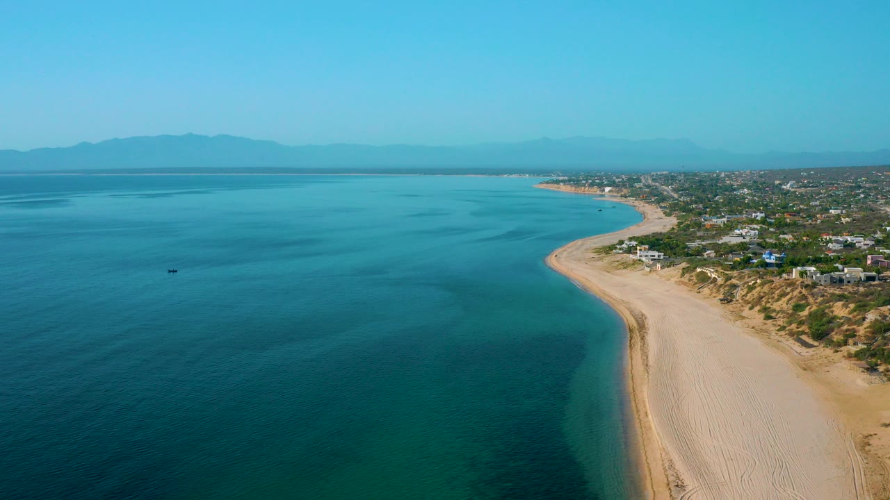 Opening aerial frame over El Sargento La Ventana, Mexico, showing turquoise water beside a wide curving sand beach, scattered coastal homes, hazy mountains, and no visible people.