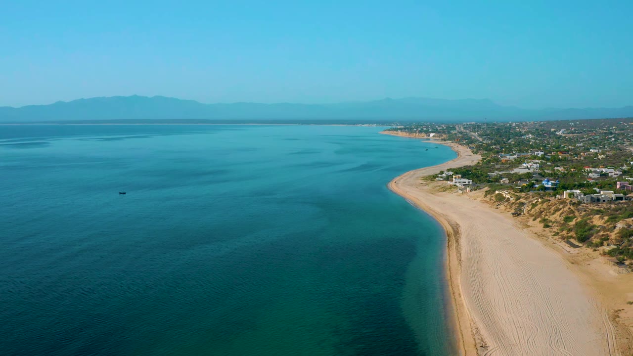 High coastal view with clear water, pale sand, small offshore boat, and low beach town.