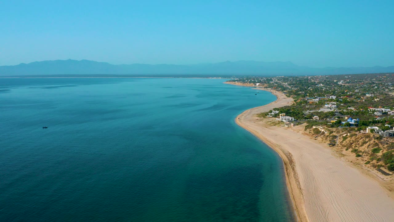 High aerial view over El Sargento La Ventana, Mexico, with blue water on the left, a sandy beach curving along the right, coastal homes beyond the dunes, distant mountains, and no visible people.