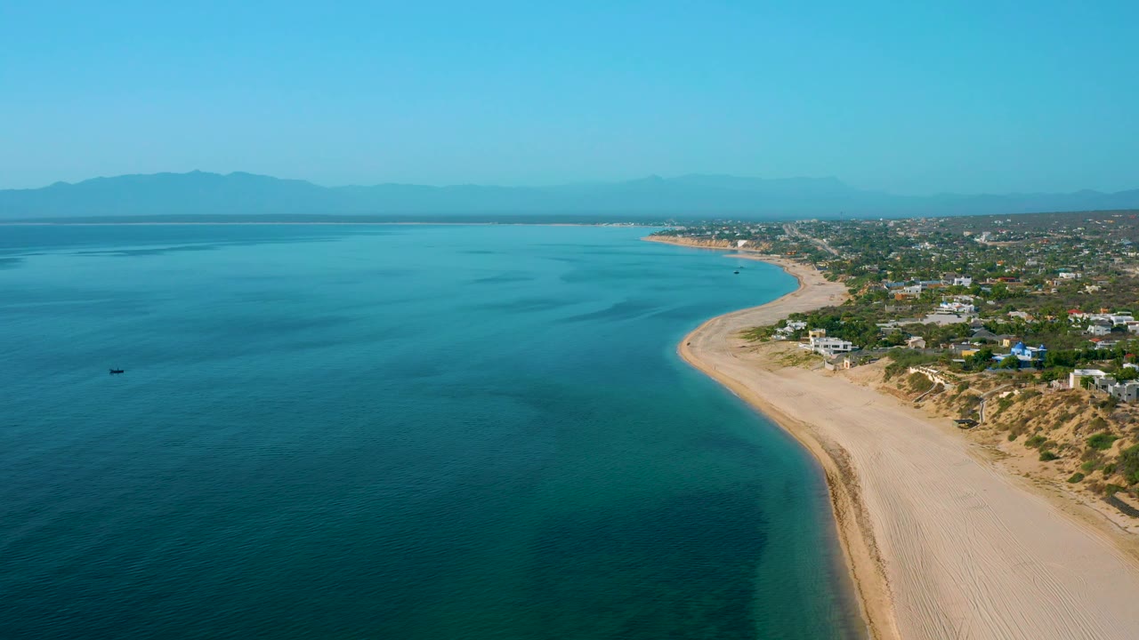 High view of El Sargento La Ventana, Mexico, with darker blue water filling more of the foreground, a pale beach strip along the right edge, low coastal buildings, mountains, and no visible people.