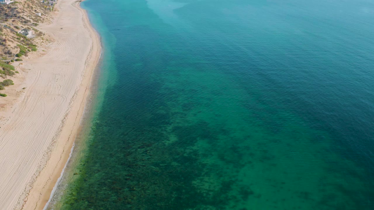 Aerial frame over El Sargento La Ventana, Mexico, with pale sand on the left, clear turquoise water on the right, dark underwater reef patches near shore, and no visible people.