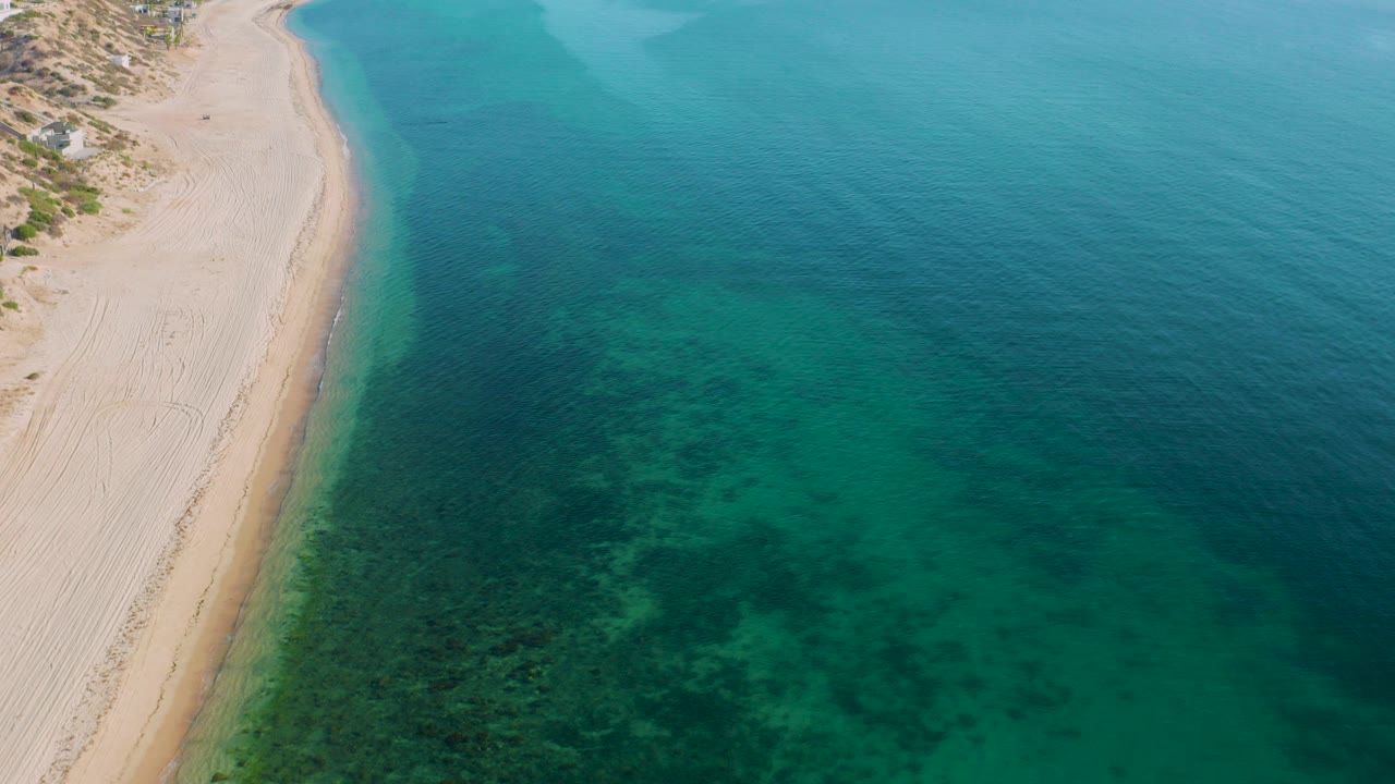 Aerial frame from El Sargento La Ventana, Mexico showing pale sand along the left side, blue-green water with dark underwater patches, and no visible people.