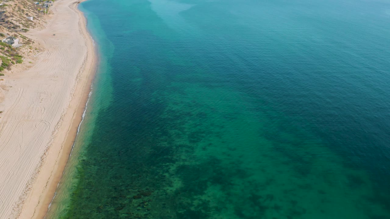 Aerial frame from El Sargento La Ventana, Mexico showing more open water to the right, sand and sparse green plants at the upper left, dark underwater areas, and no visible people.
