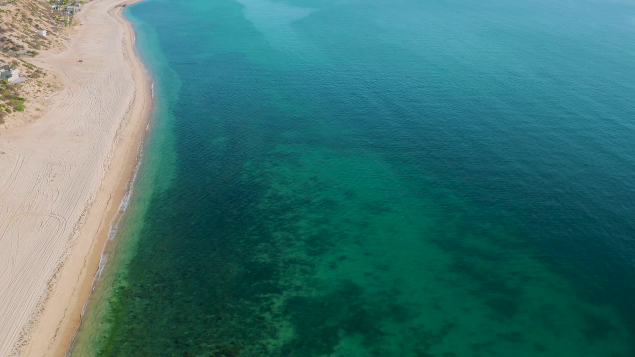 Aerial frame from El Sargento La Ventana, Mexico showing a pale beach on the left, mottled green-blue water across the center and right, faint shore wash, and no visible people.