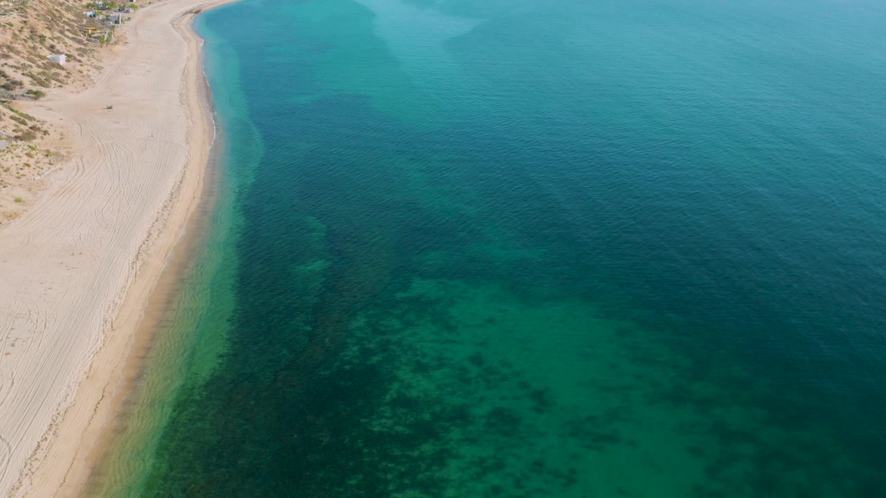 Aerial frame from El Sargento La Ventana, Mexico, showing pale sand along the left edge and turquoise water with dark underwater patches, with no visible people.