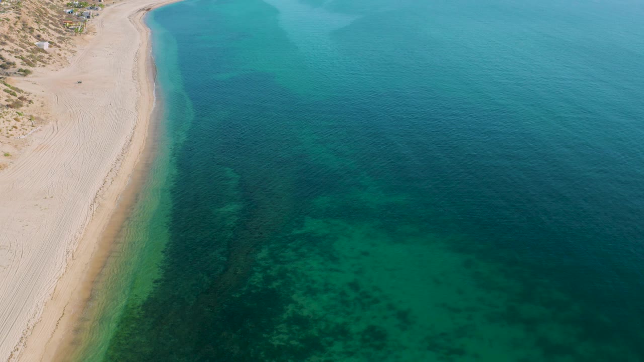 High aerial frame from El Sargento La Ventana, Mexico, showing a narrow sand strip at left, shallow green water near shore, and darker blue sea to the right, with no visible people.