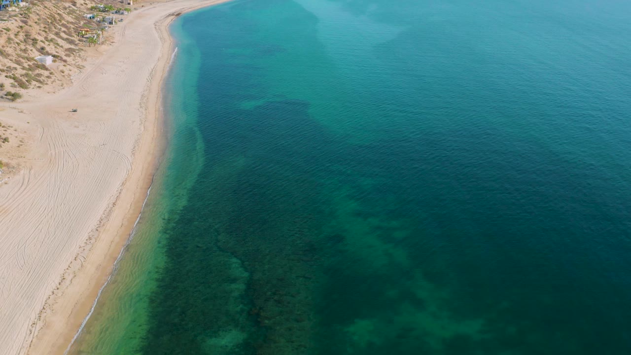 Aerial view in El Sargento La Ventana, Mexico, showing the beach running diagonally beside calm shallow water, dune texture at upper left, and no visible people.