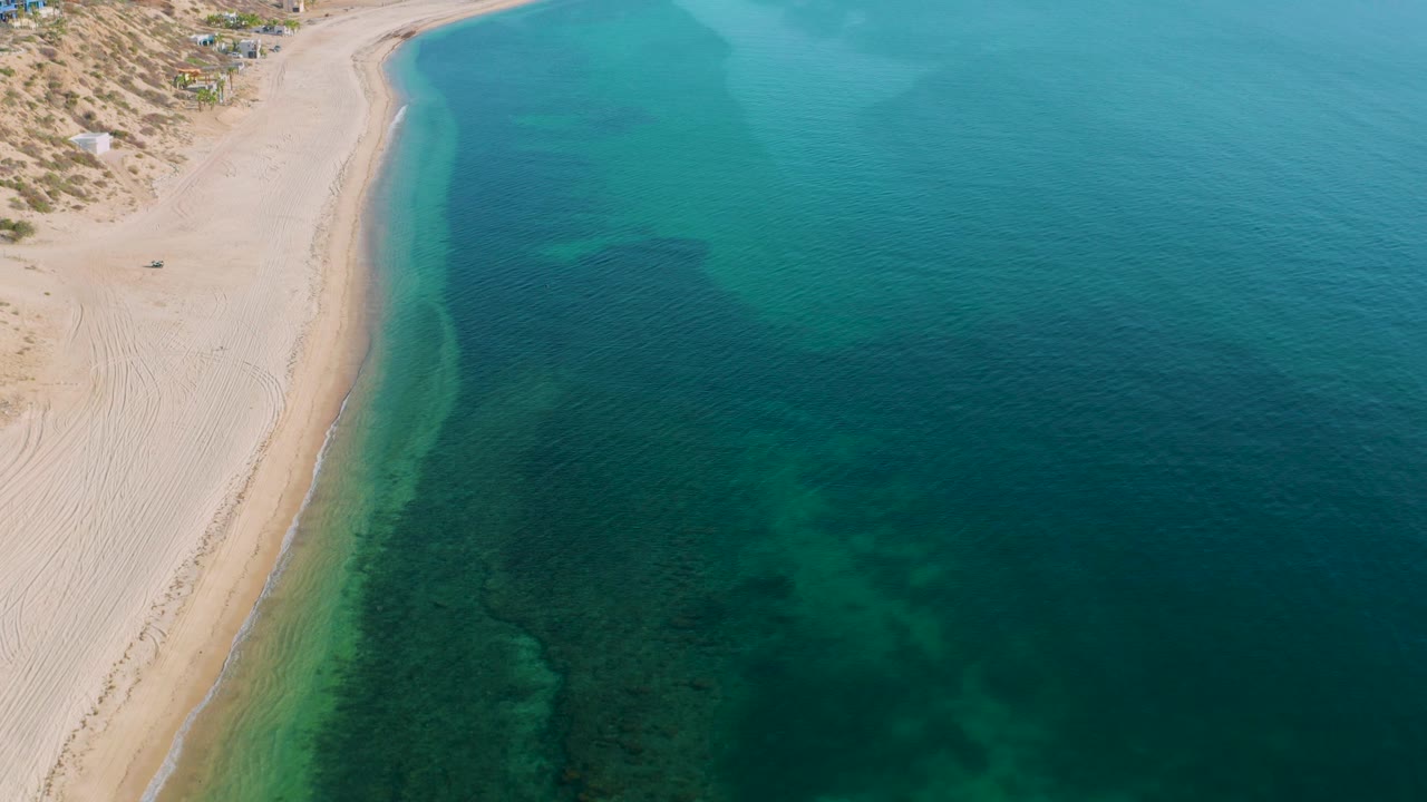 High drone-like view over El Sargento La Ventana, Mexico, with the empty beach along the left side, turquoise water spreading across the frame, soft wave lines, and no visible people.