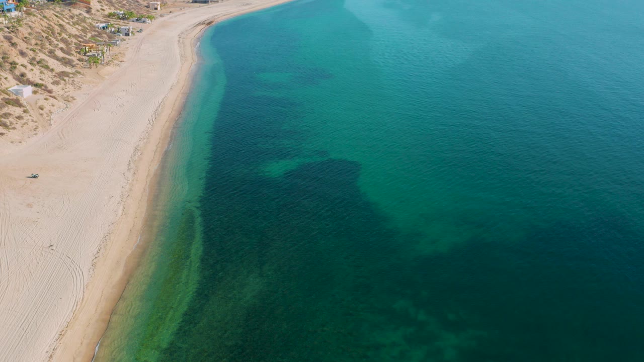 Opening aerial frame over El Sargento La Ventana, Mexico, with pale sand curving along clear green water, dark underwater patches offshore, dune vegetation inland, and no visible people.