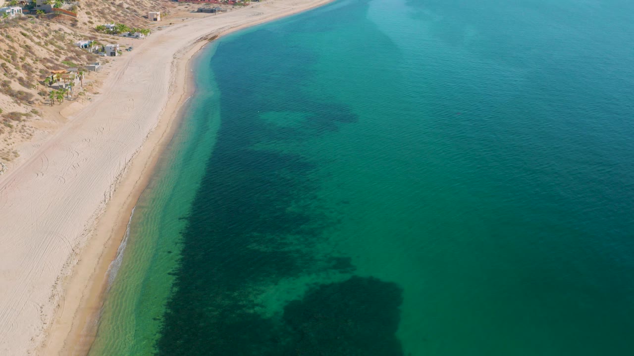 Opening aerial frame over El Sargento La Ventana, Mexico, with pale sand on the left, turquoise green water on the right, dark underwater patches near shore, scattered coastal buildings inland, and no visible people.