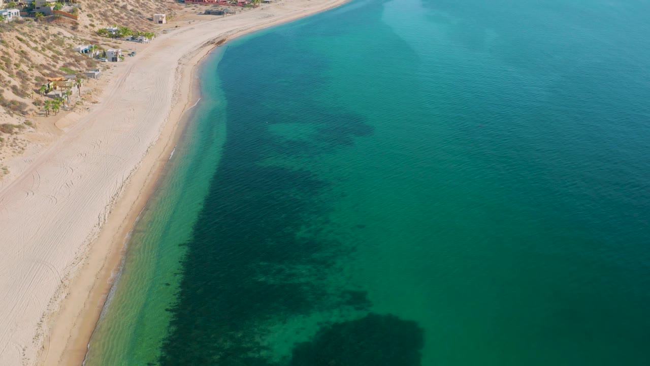 Aerial frame over El Sargento La Ventana, Mexico, with the sandy beach narrowing along the left, broad turquoise water filling the frame, dark submerged areas close to shore, and no visible people.