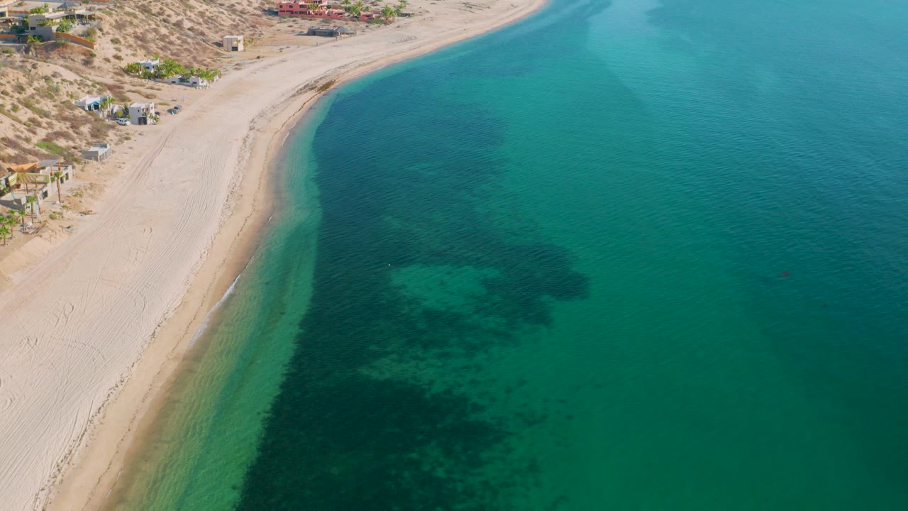 Aerial frame over El Sargento La Ventana, Mexico, showing a curved pale beach, turquoise shallows, darker underwater plant patches, dry hillside terrain, scattered buildings, and no visible people.