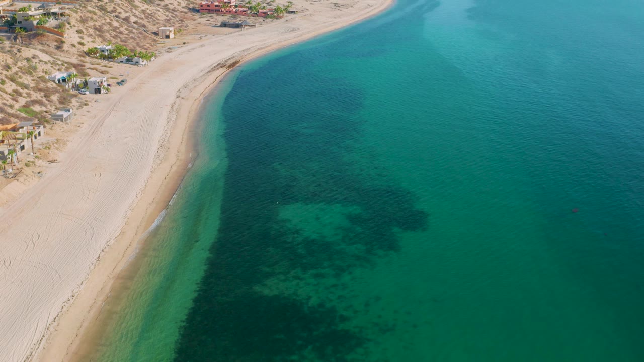 Aerial coastline view over El Sargento La Ventana, Mexico, showing pale sand along the left side, broad turquoise sea on the right, darker underwater growth, sparse buildings above the beach, and no visible people.