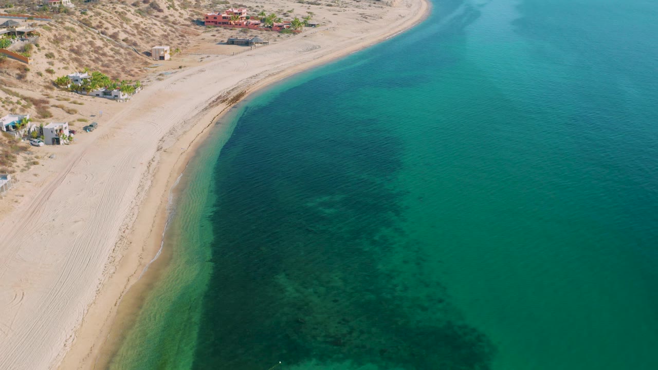 Aerial frame over El Sargento La Ventana, Mexico, showing the beach curve closer to center, dark seaweed-like patches under blue-green water, desert vegetation inland, and no visible people.